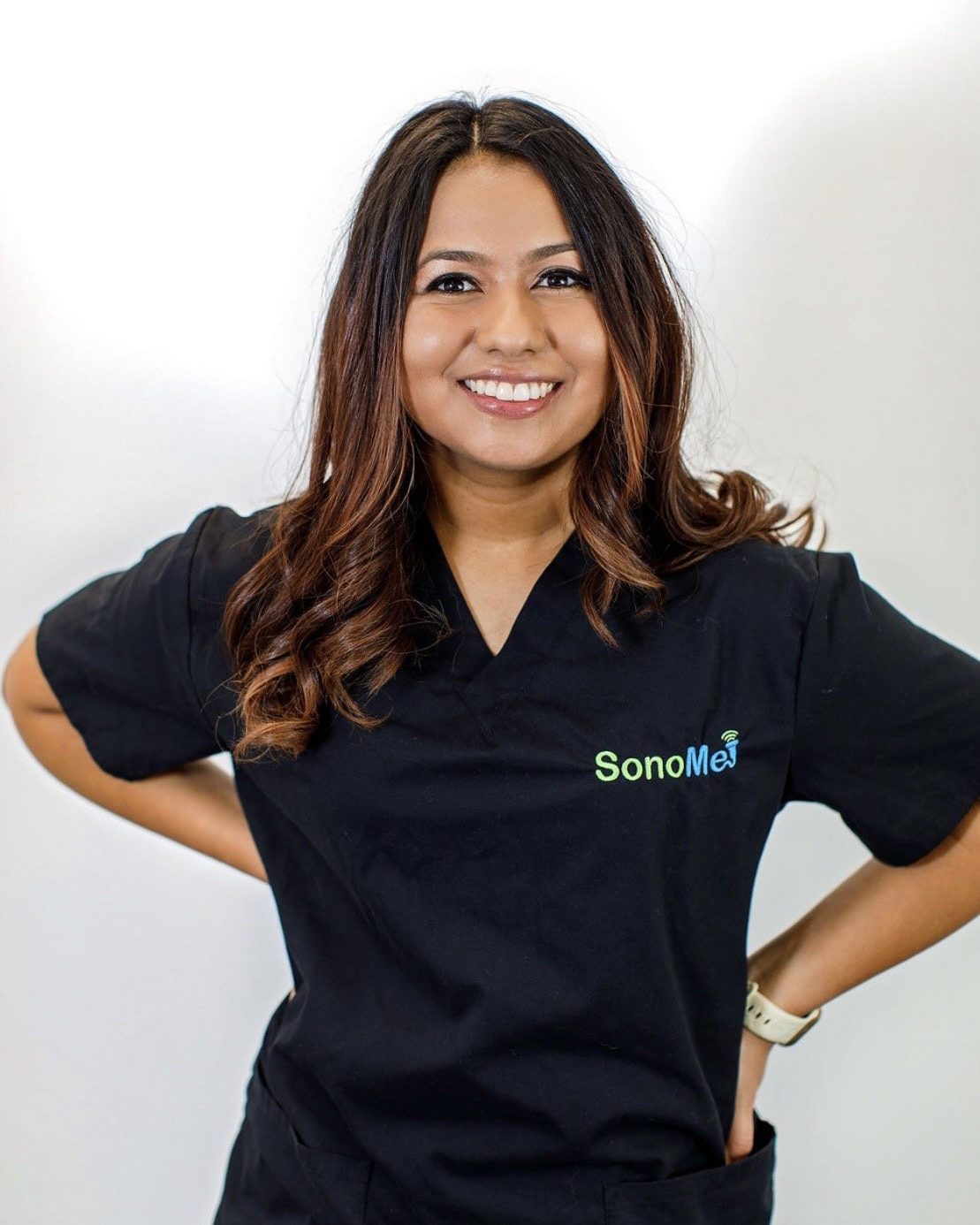 A woman smiling and posing with her hands on her hips, wearing black scrubs with the logo 'SonoMe' on it.